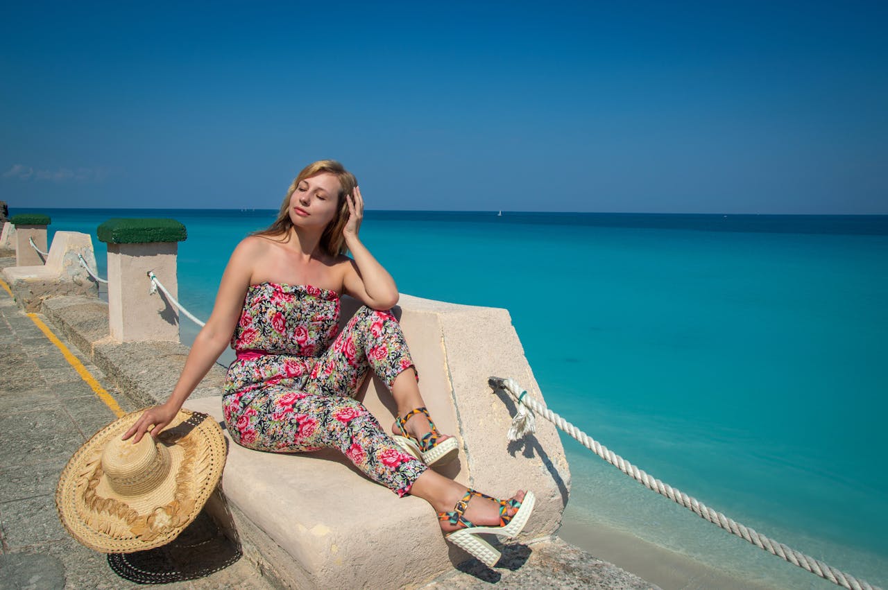 A young woman in a floral jumpsuit relaxes by the turquoise ocean on a sunny day, exuding summer vibes.