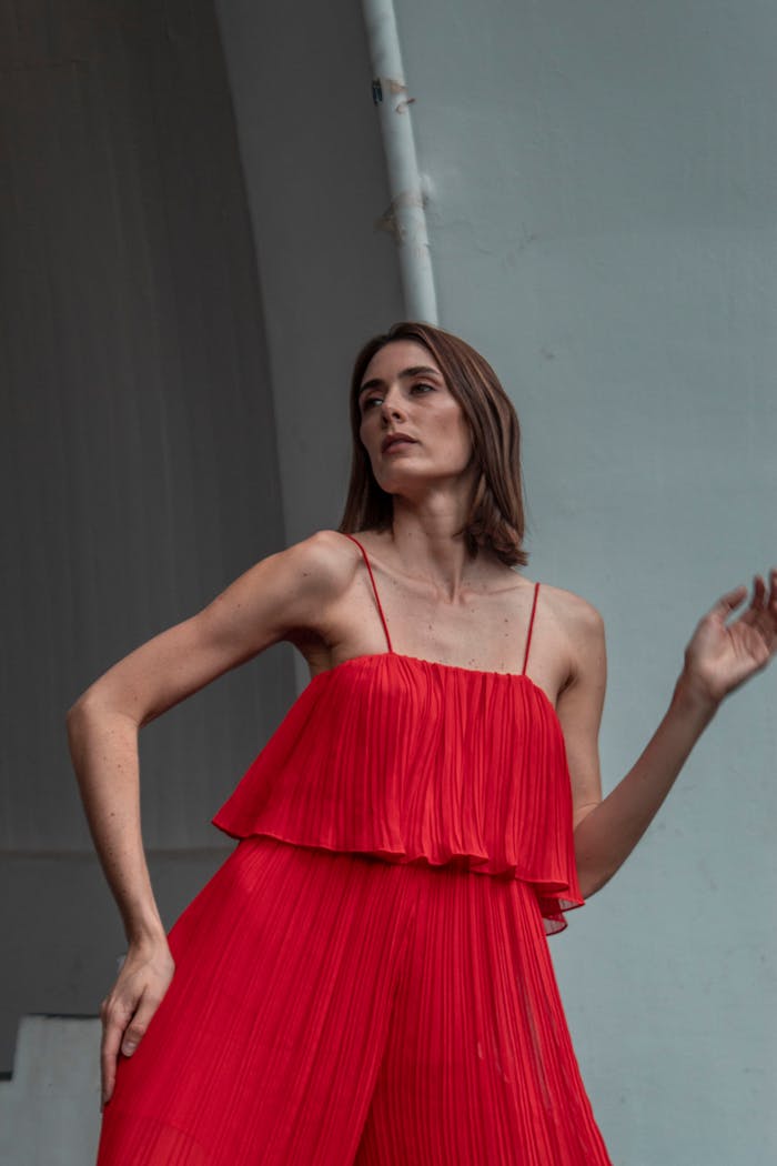 Stylish young woman wearing a red jumpsuit poses elegantly outdoors against an urban backdrop.